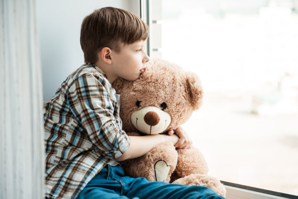 Trauriger Junge sitzt am Fenster und hält seinen Teddybären